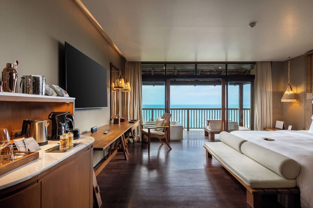 A hotel room with a large bed, a wooden desk, a TV, and a balcony with a view of the sea.