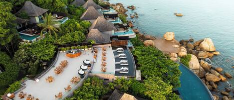 An aerial view of a resort with multiple thatched-roof villas, a central pool area with lounge chairs, and a rocky coastline.