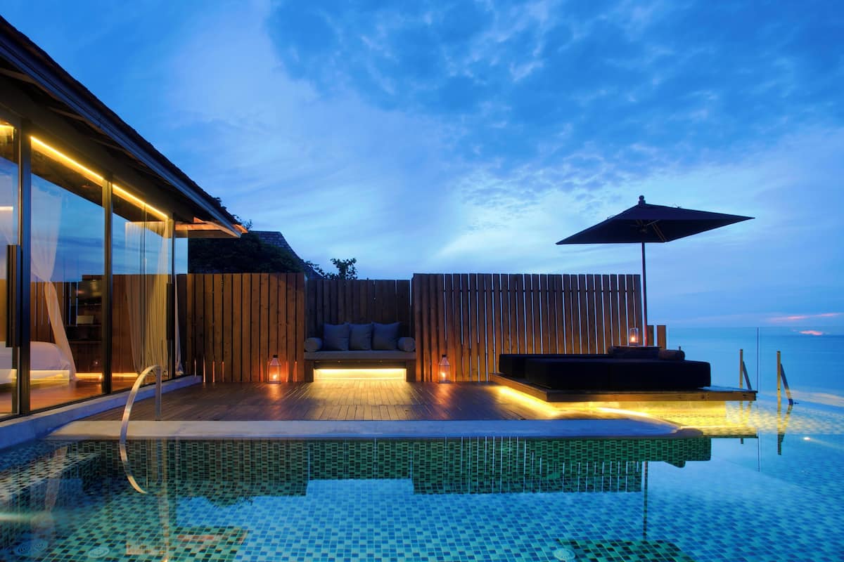 A modern infinity pool with a glass edge, a wooden deck, and a seating area with cushions and a table.