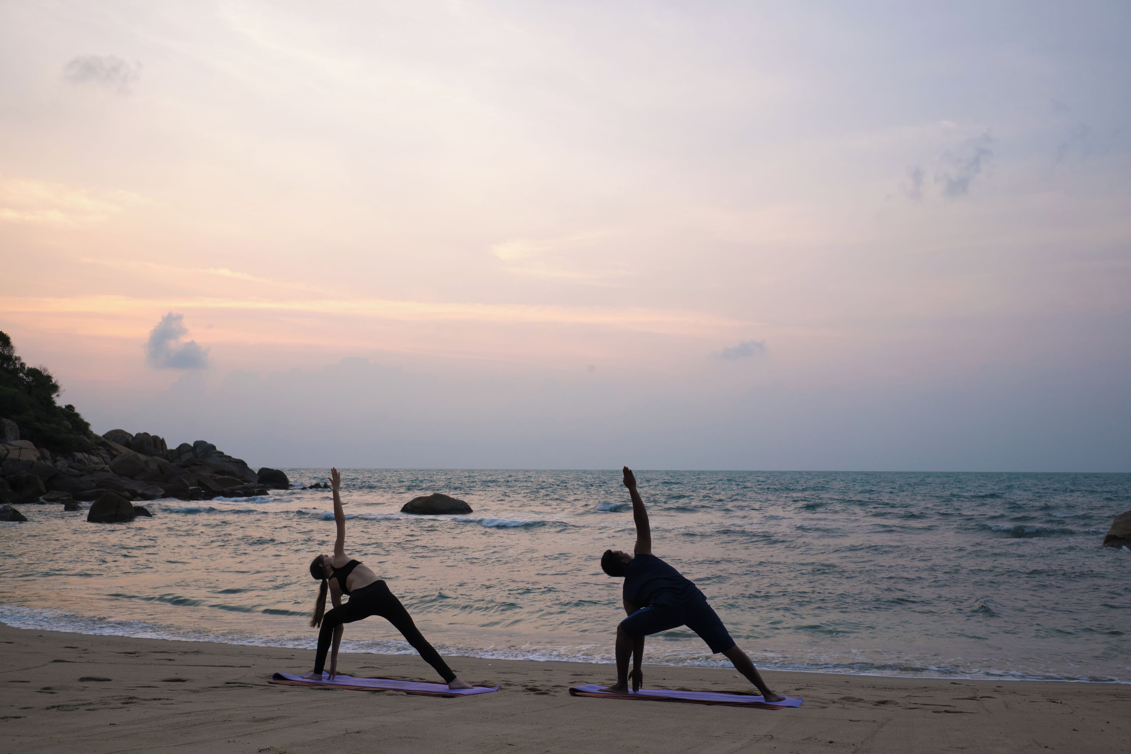 Two individuals practicing yoga on mats at a beach with a rocky shoreline and ocean in the background.
