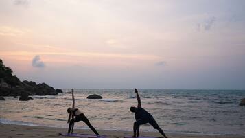 Two individuals practicing yoga on mats at a beach with a rocky shoreline and ocean in the background.