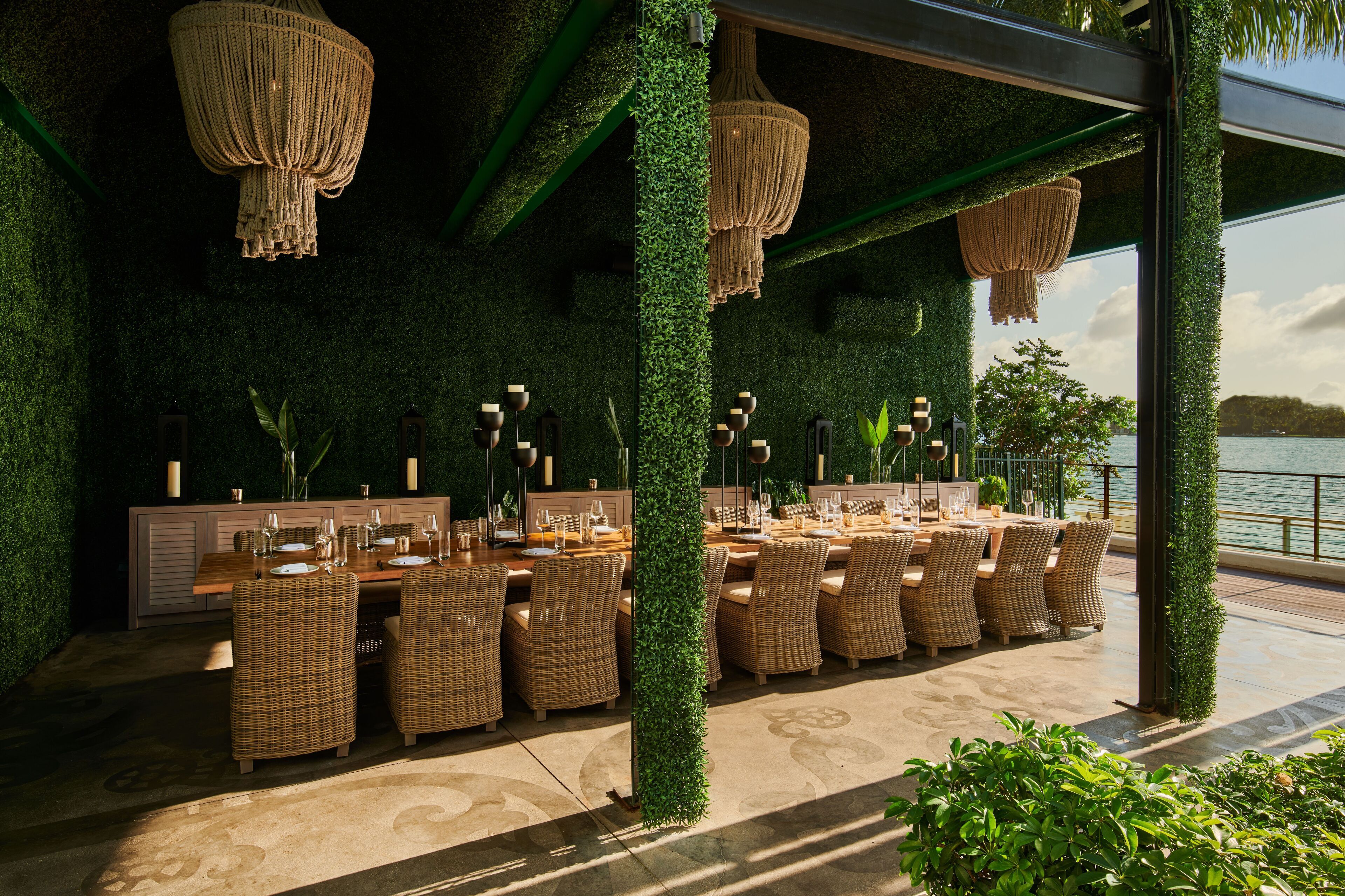 An outdoor dining area with wicker chairs and tables, surrounded by greenery and a view of the water.