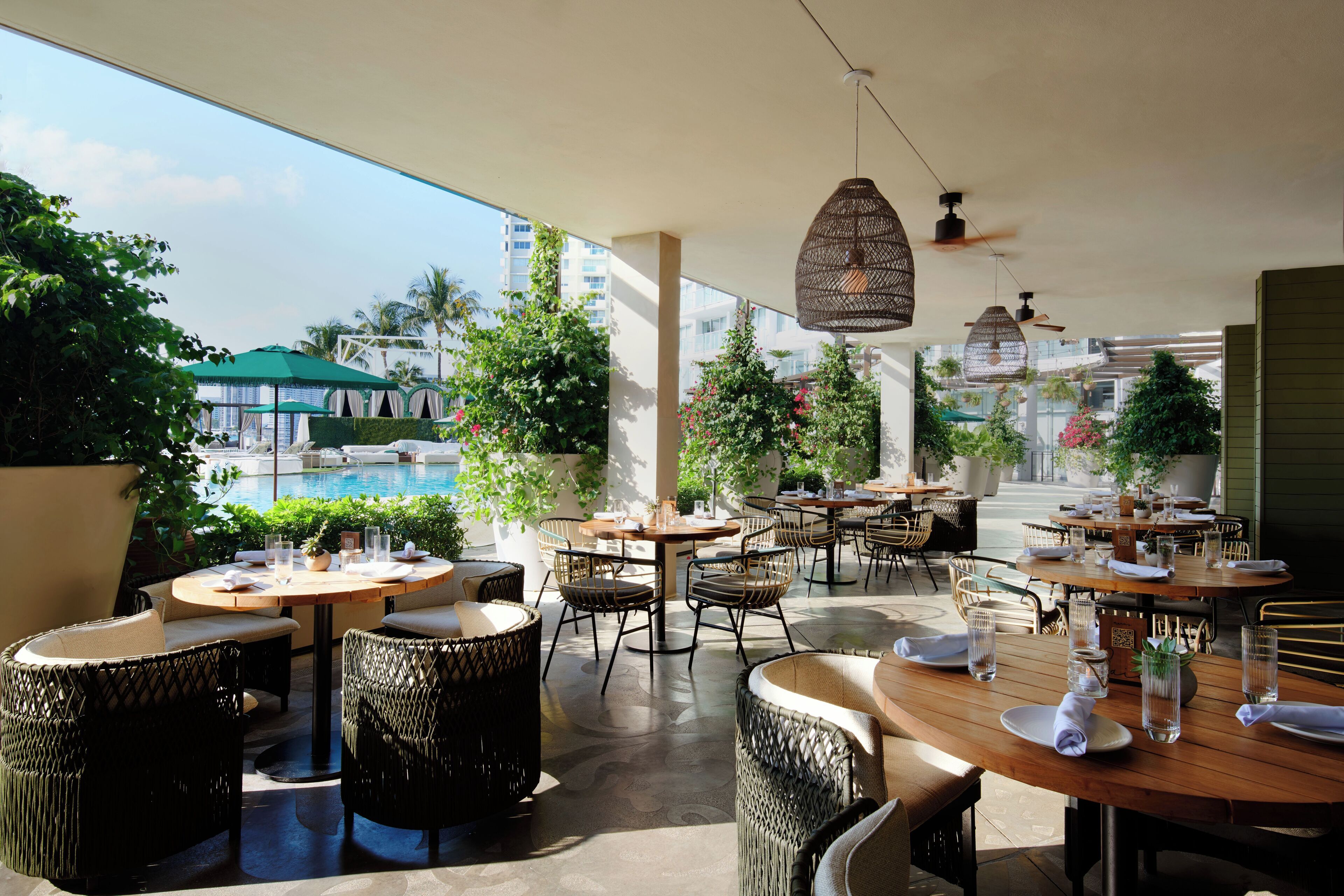 An outdoor dining area with wicker furniture and tables, surrounded by greenery and a view of a pool and buildings.