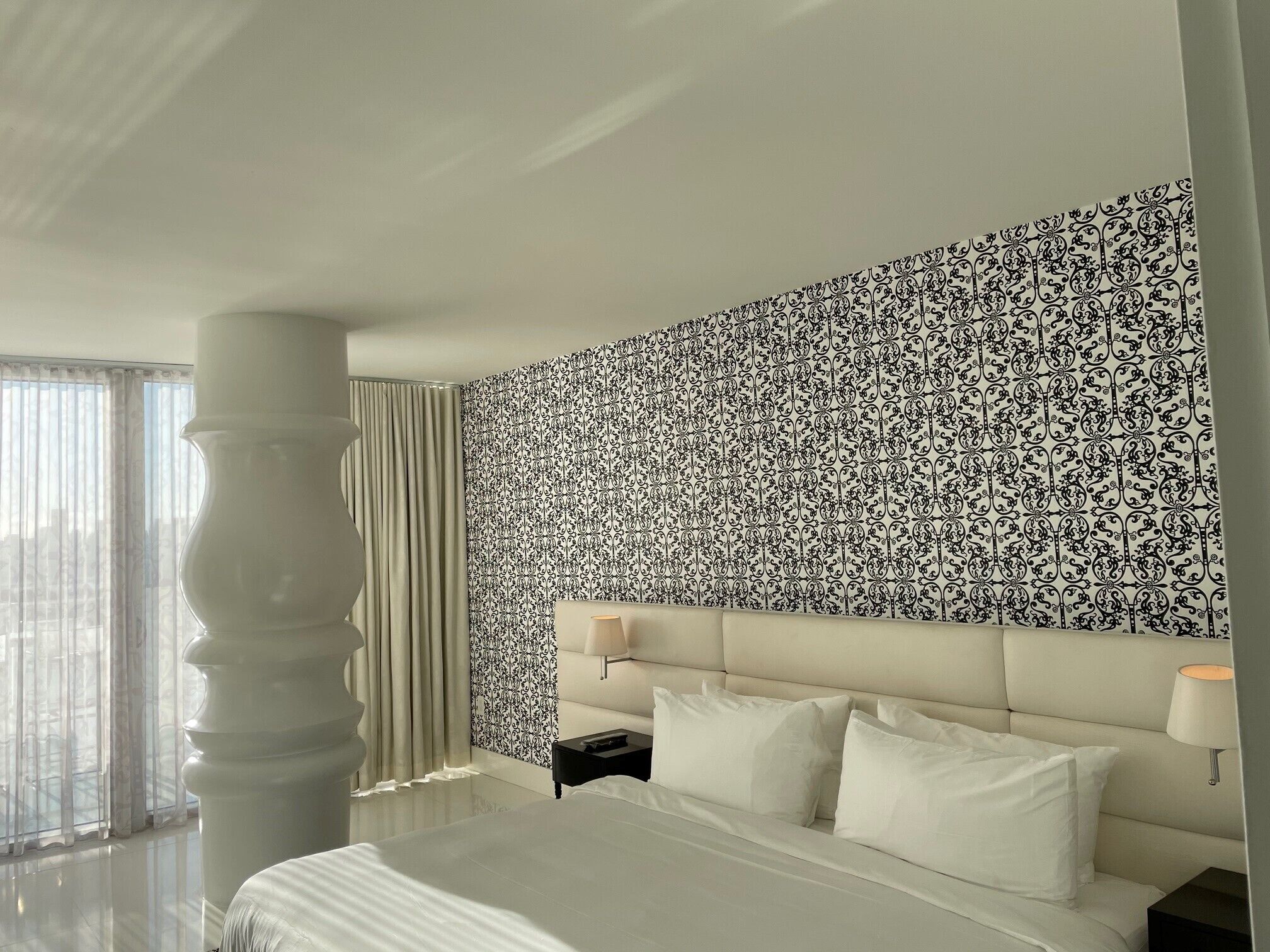 A hotel room with a large bed, a headboard with a light-colored upholstered headboard, a nightstand with a lamp, and a patterned wallpaper on the wall.