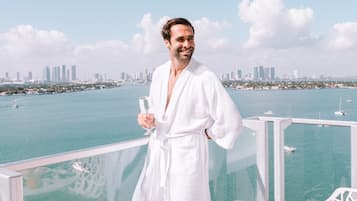A person in a white robe standing on a balcony with a table of breakfast items and a glass of orange juice.