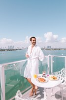 A person in a white robe standing on a balcony with a table of breakfast items and a glass of orange juice.