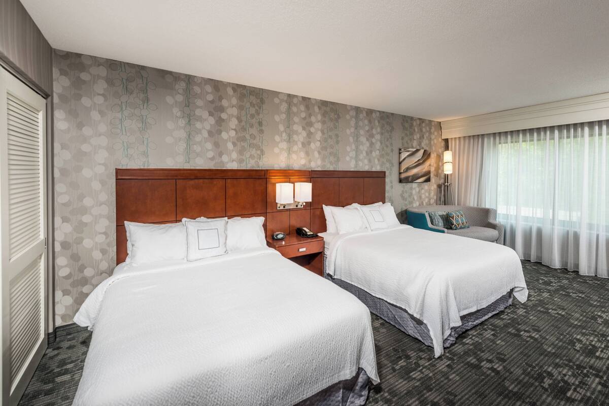 A hotel room with two beds, a wooden headboard, a nightstand with a lamp, and a view of the outdoors through a window with sheer curtains.