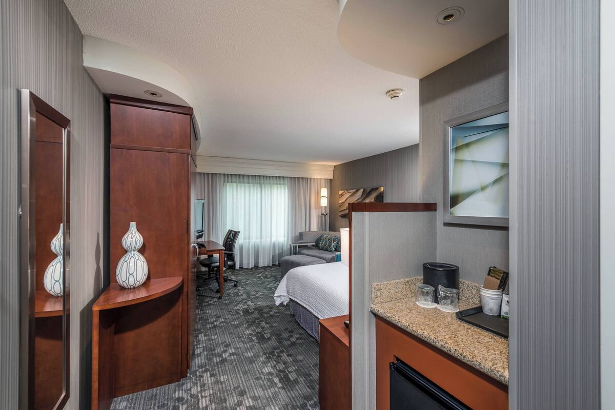 A hotel room with a bed, a desk, a chair, a vanity with a mirror, and a window with curtains.