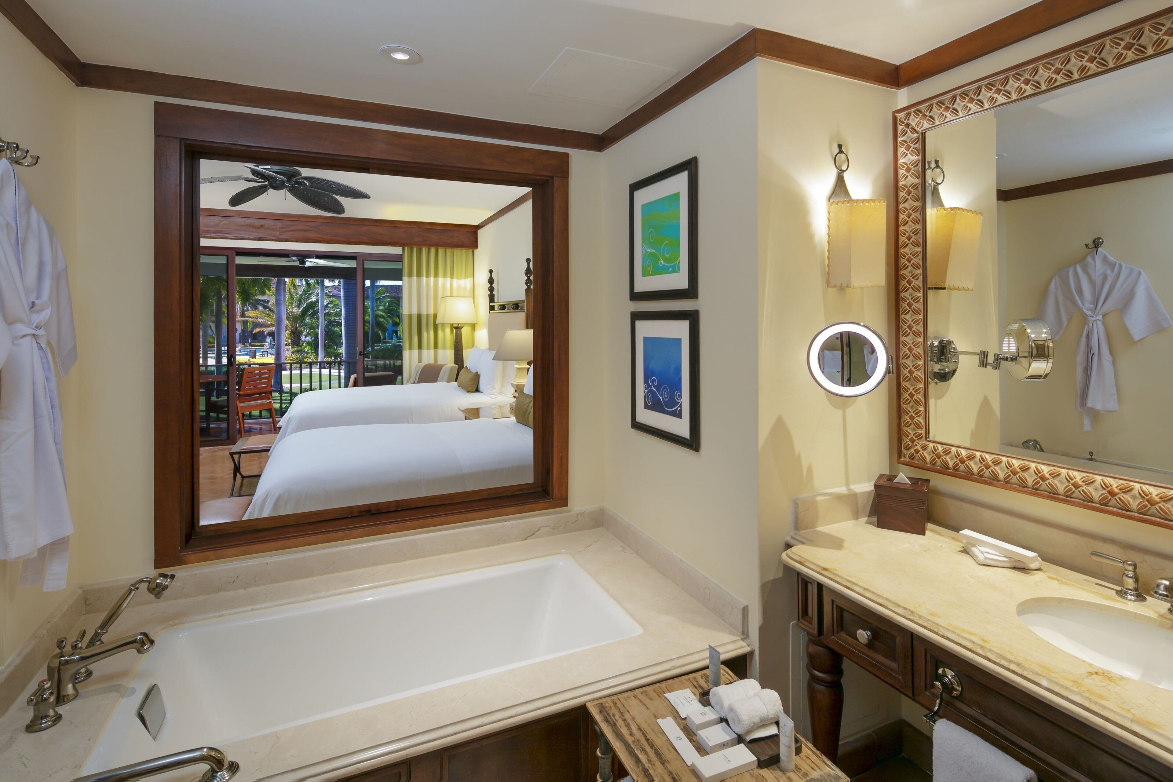 A hotel bathroom with a large bathtub, a vanity with a mirror, and a bed visible through a window.
