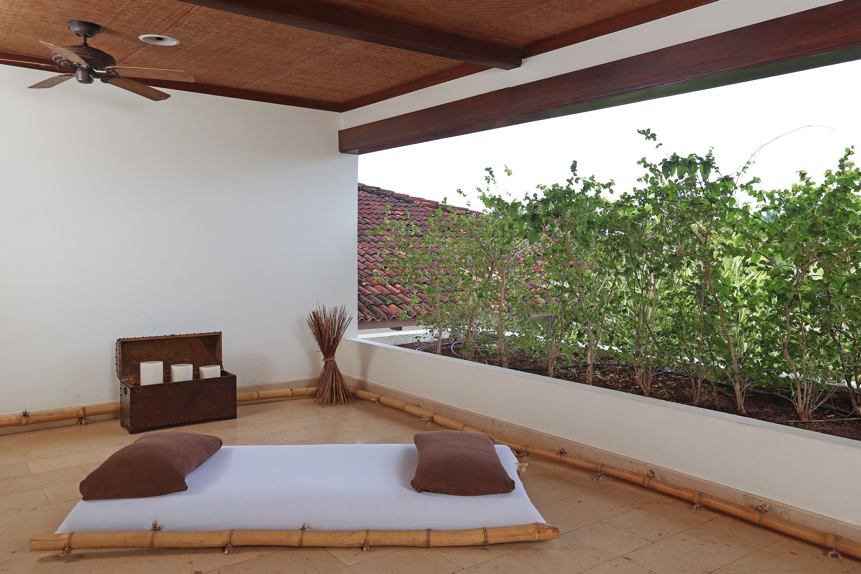 A room with a ceiling fan, a bed with two pillows, a wooden chest, and a view of trees and a building.