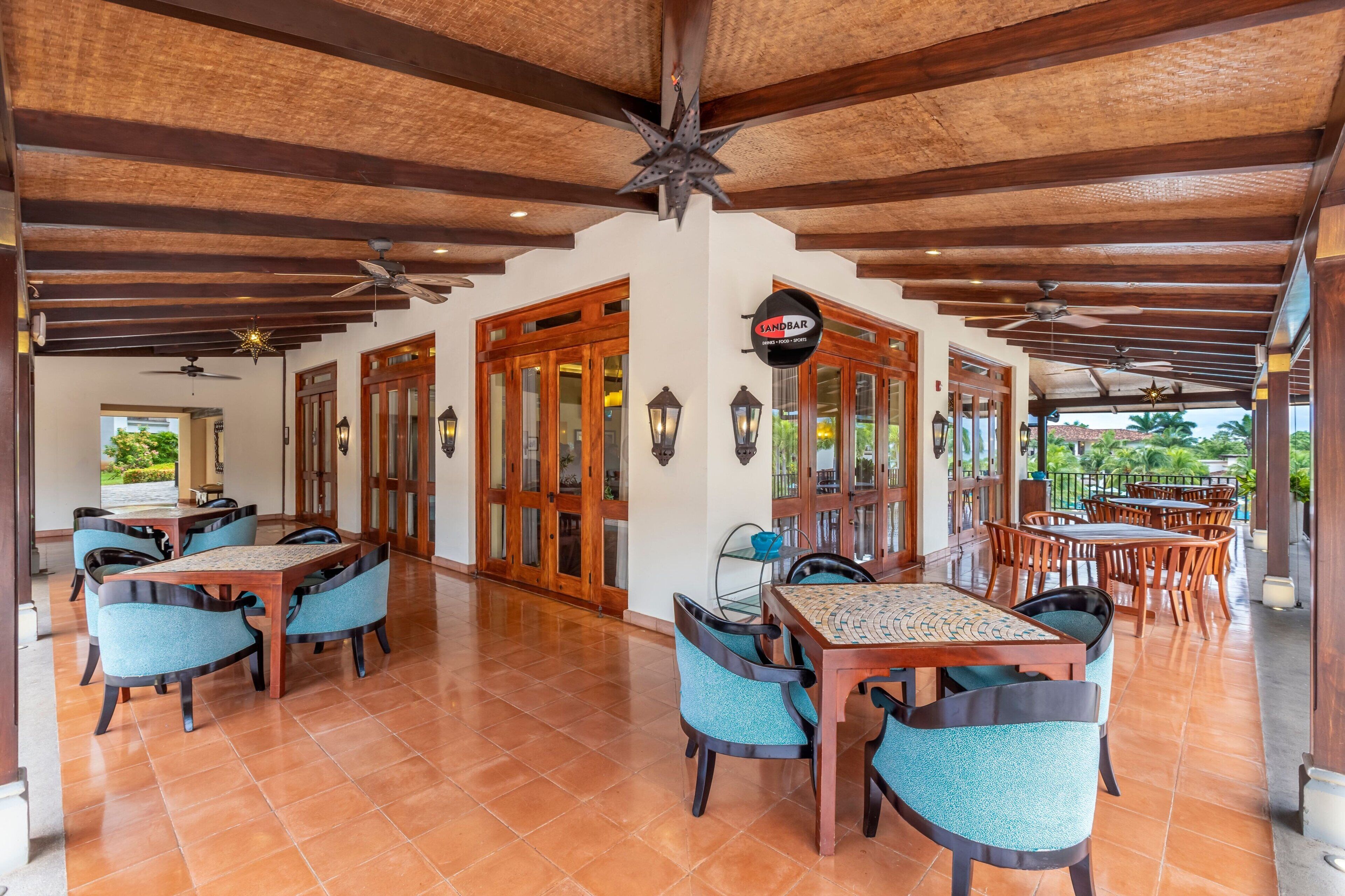 Un área de comedor al aire libre cubierta con mesas y sillas de madera, un letrero que dice 'Sandbar' y vistas del exterior.