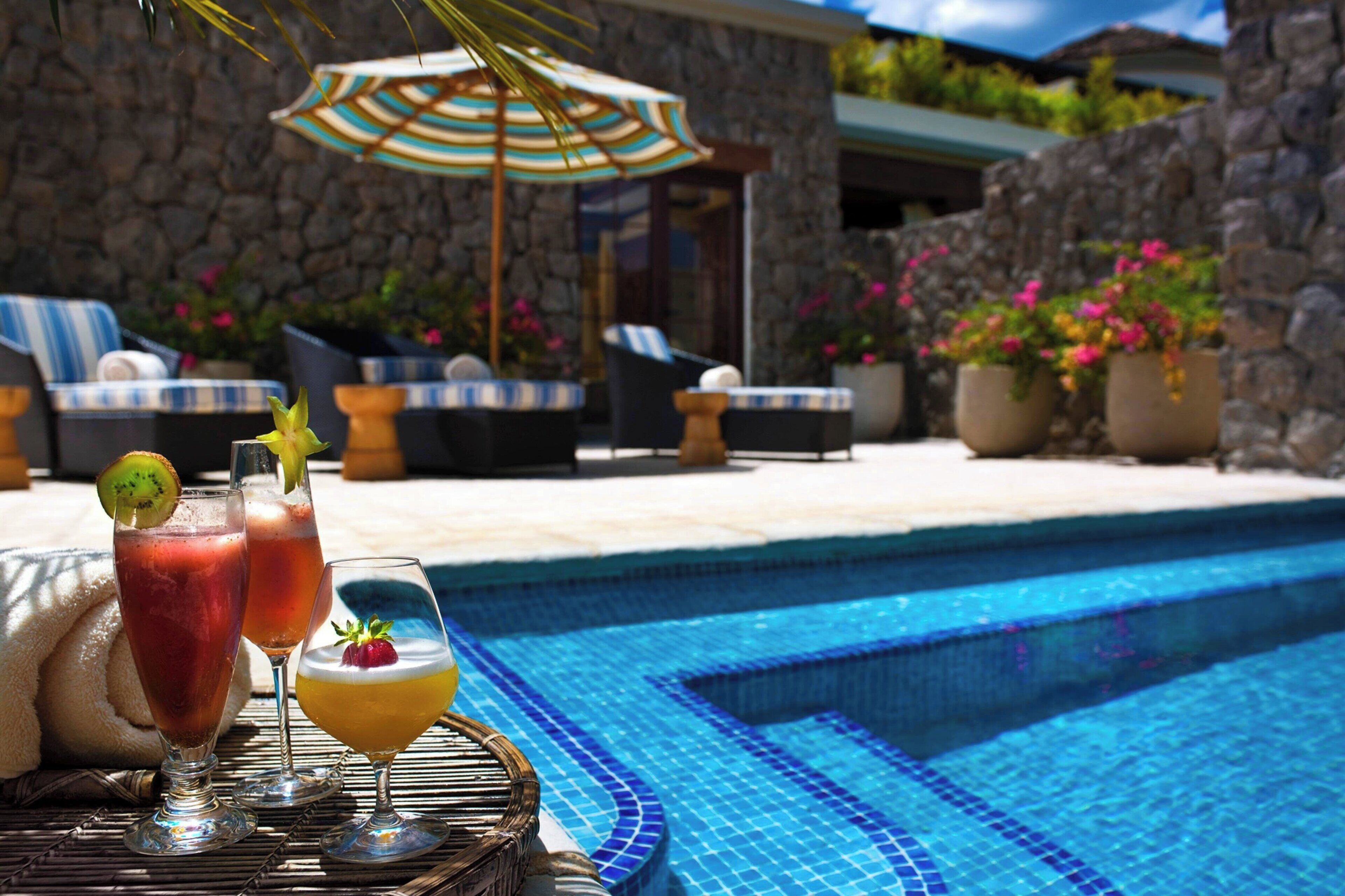 A poolside area with a stone wall, lounge chairs, and a table with drinks.