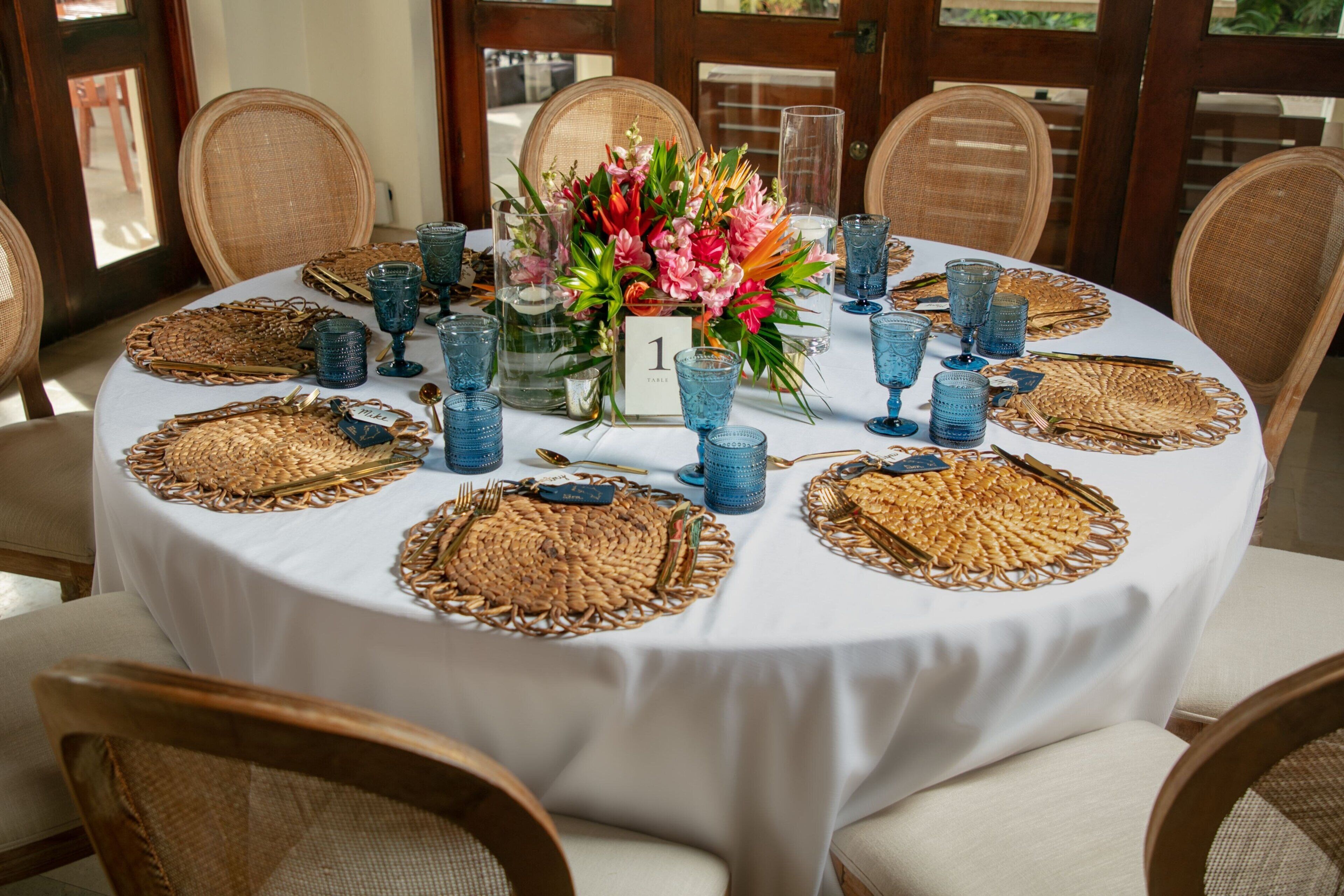 A round table with a white tablecloth, blue glassware, and woven placemats.