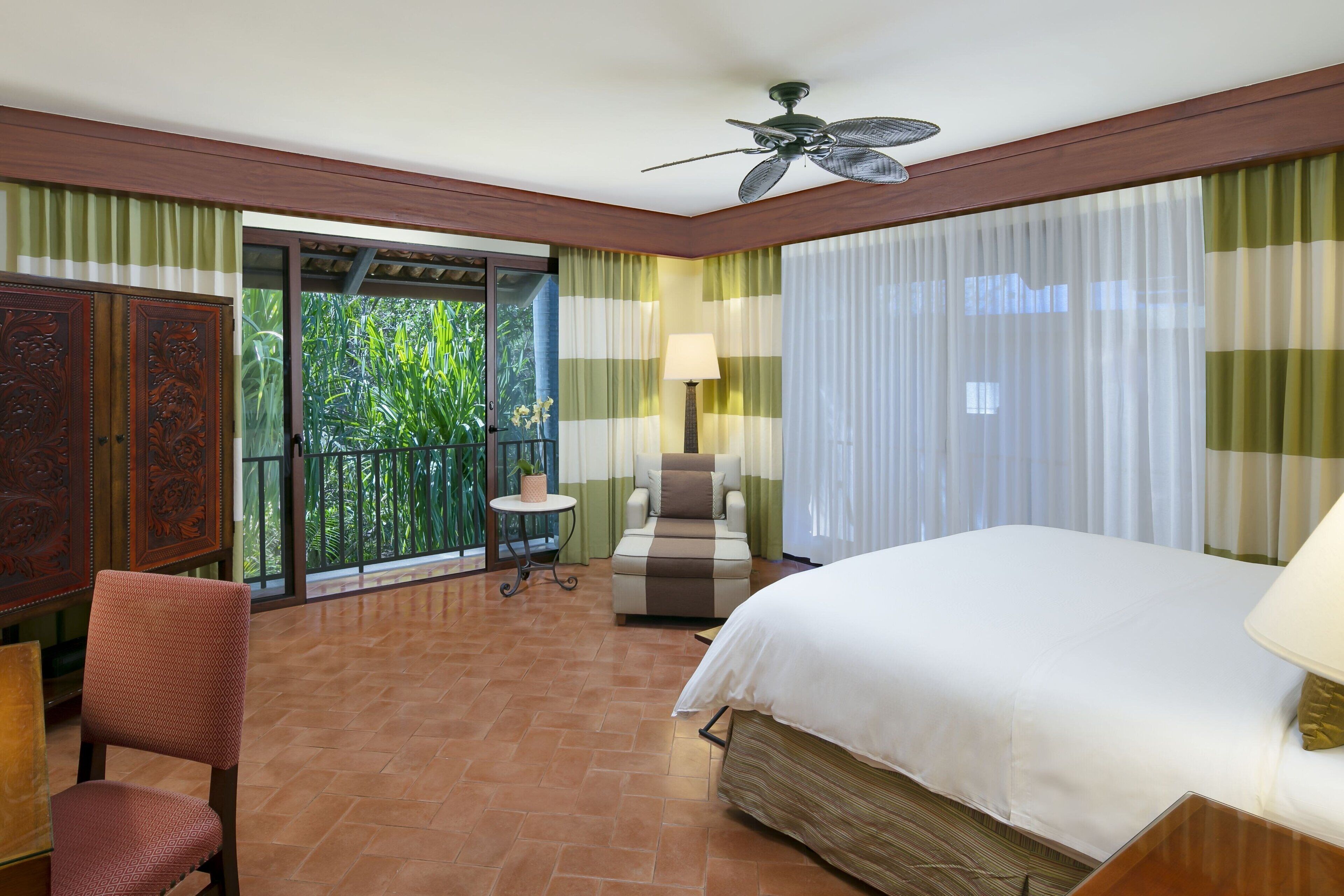 A hotel room with a bed, a chair, a table, and a balcony with a view of greenery.