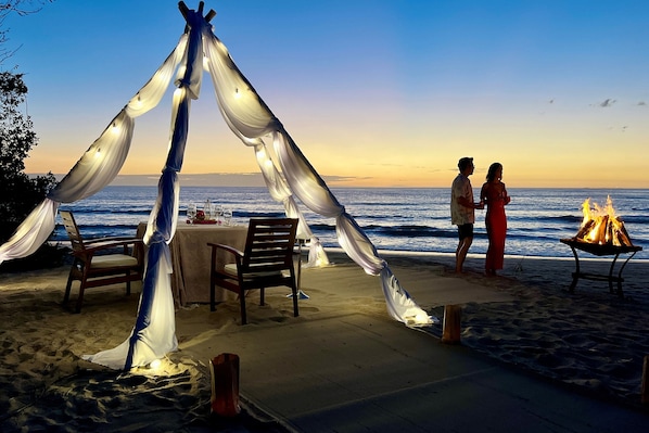 Un'area pranzo in spiaggia con tavolo e sedie, un braciere e una struttura simile a una tenda con tessuto drappeggiato.