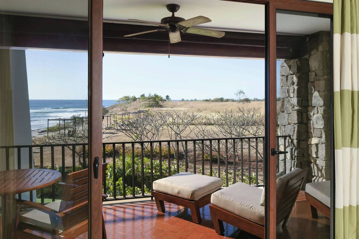 A balcony with a view of the ocean, a wooden table, and two cushioned chairs.