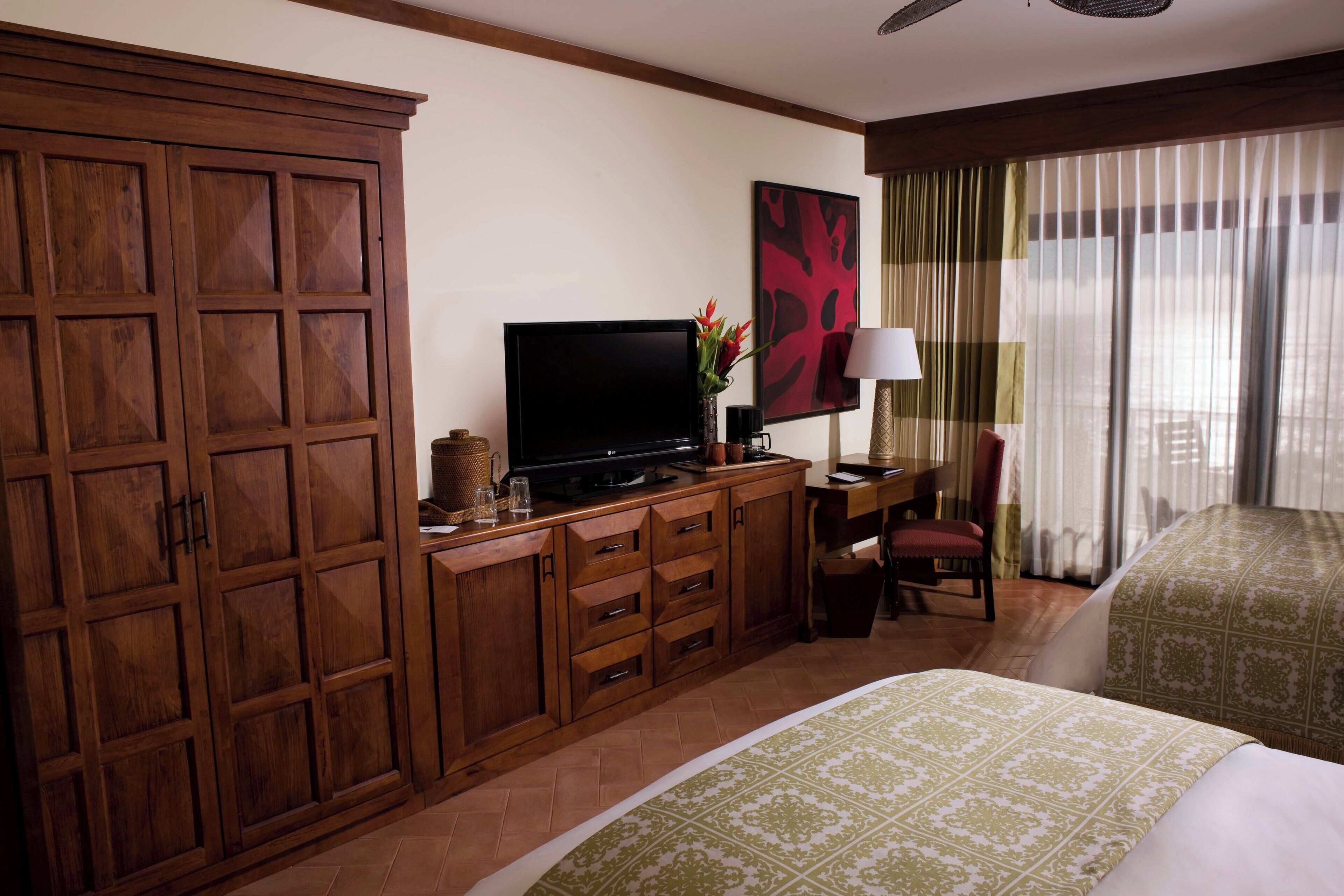 Ein Hotelzimmer mit großem Holzschrank, Flachbildfernseher, Schreibtisch mit Lampe, Stuhl und einem Bett mit gemusterten Betttextilien.