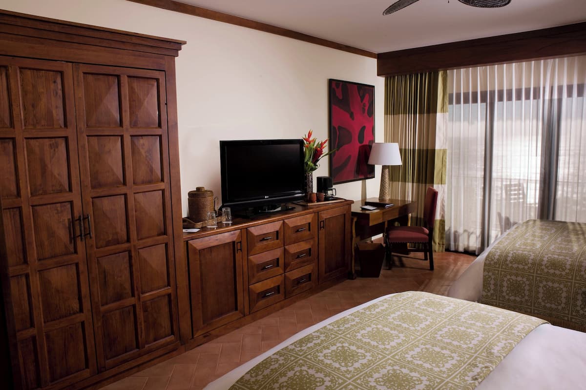 Ein Hotelzimmer mit großem Holzschrank, Flachbildfernseher, Schreibtisch mit Lampe, Stuhl und einem Bett mit gemusterten Betttextilien.