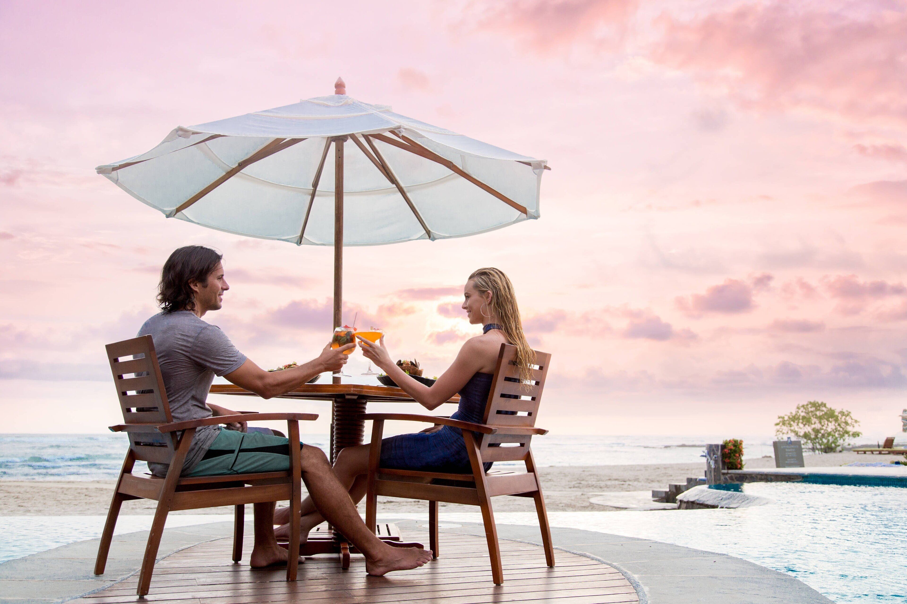 Una pareja cenando en una mesa con sombrilla, con vista a una playa y una piscina.