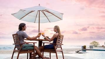 Una pareja cenando en una mesa con sombrilla, con vista a una playa y una piscina.