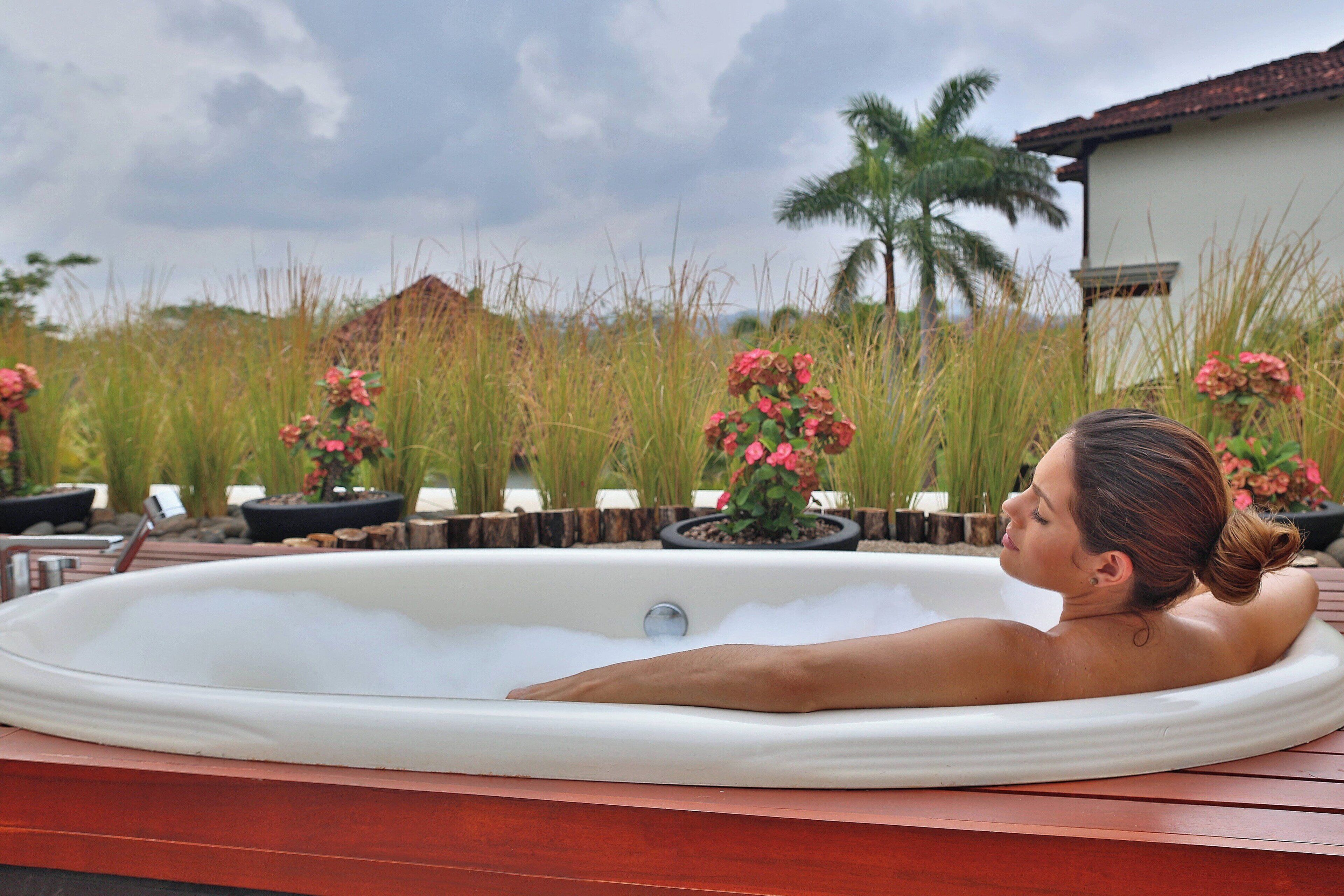A hot tub with a woman relaxing, surrounded by potted plants and a palm tree.