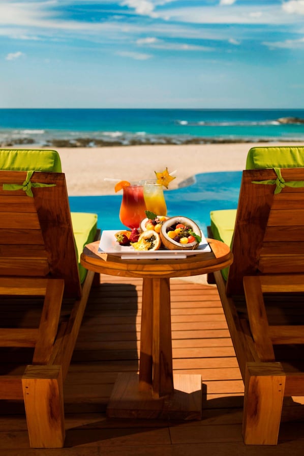 Una terraza de madera con dos sillas verdes acolchadas mirando a una playa, con una mesa en el centro que sostiene un plato de fruta, una jarra de jugo y una decoración con forma de estrella.