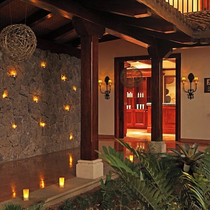 A stone wall with recessed lighting, a wooden ceiling, and a doorway leading to a room with a red door.