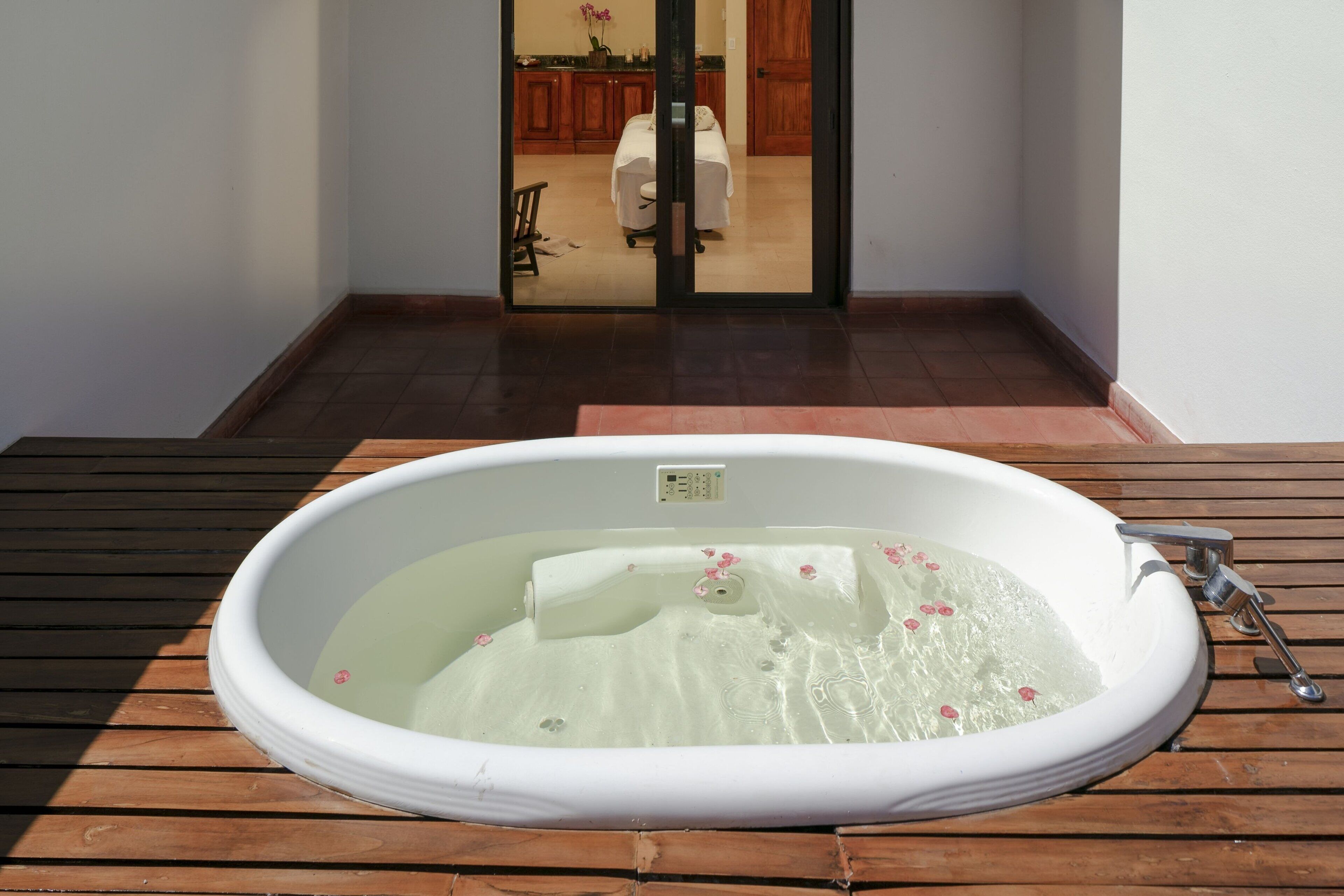 A hot tub on a wooden deck with a view of a room with a bed and a cabinet in the background.