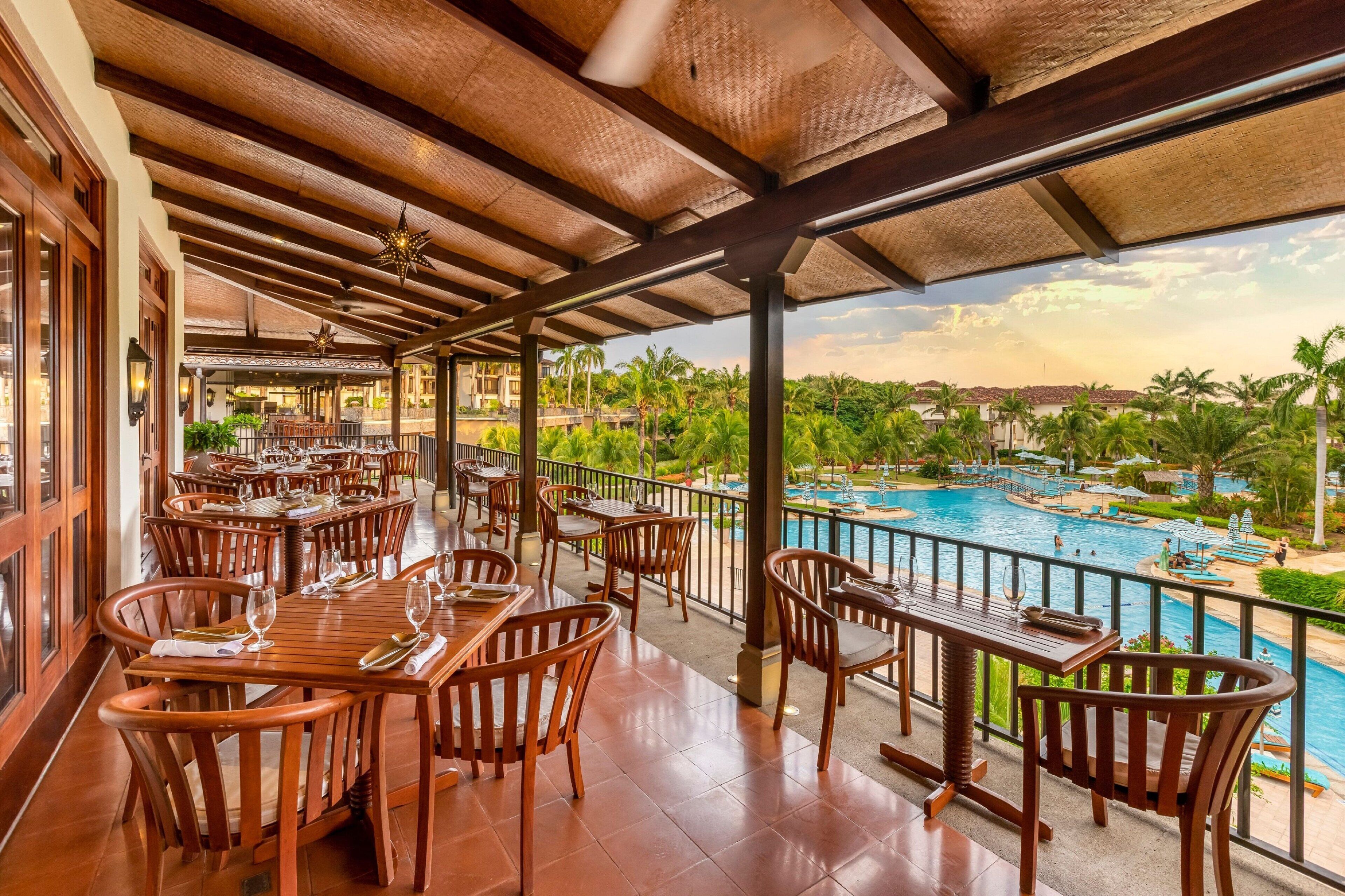 A restaurant with wooden tables and chairs, a pool with lounge chairs, and a view of palm trees and a mountain range.