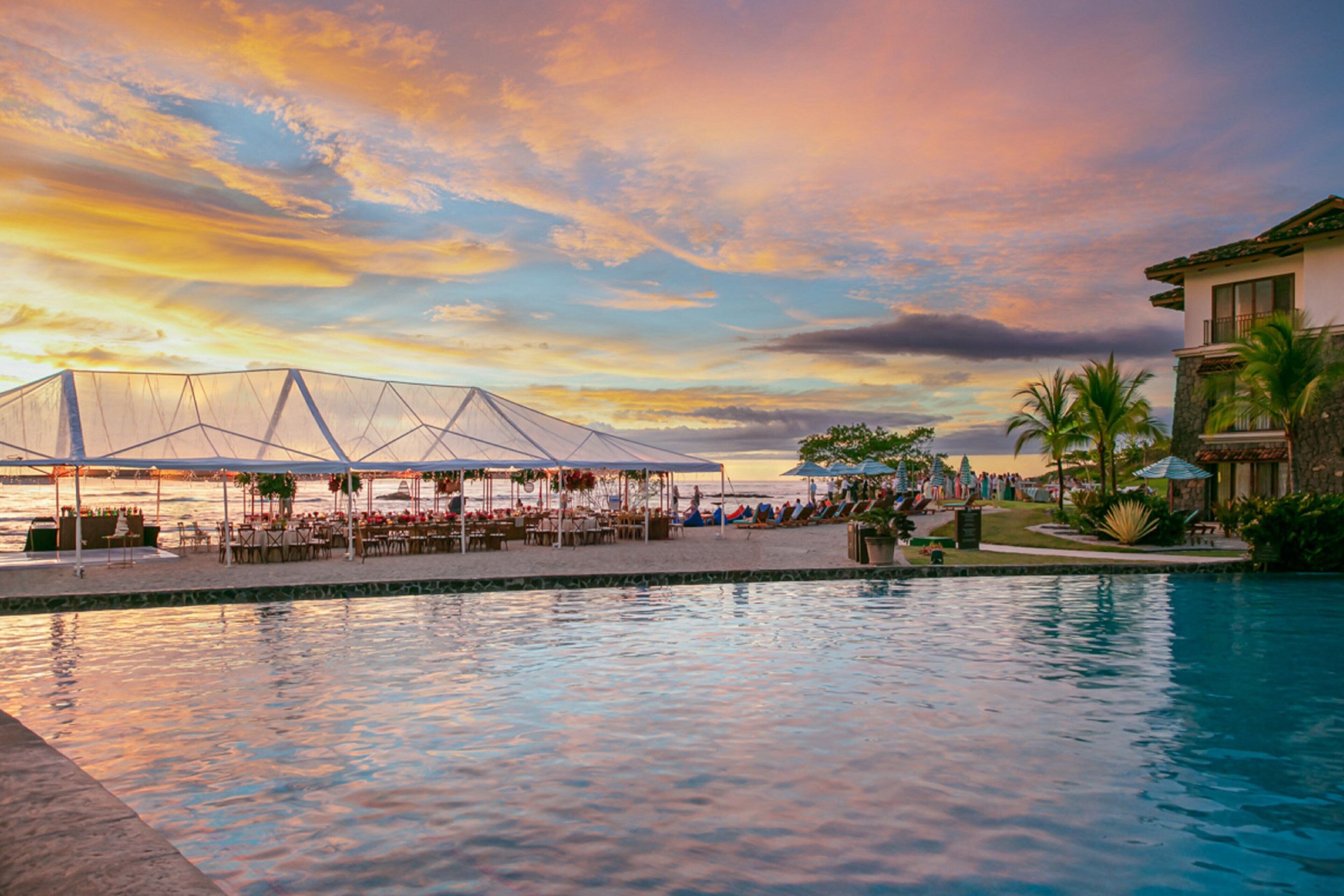 Un espace aménagé au bord de la piscine, avec un coin repas couvert et une vue sur l’océan au coucher du soleil.