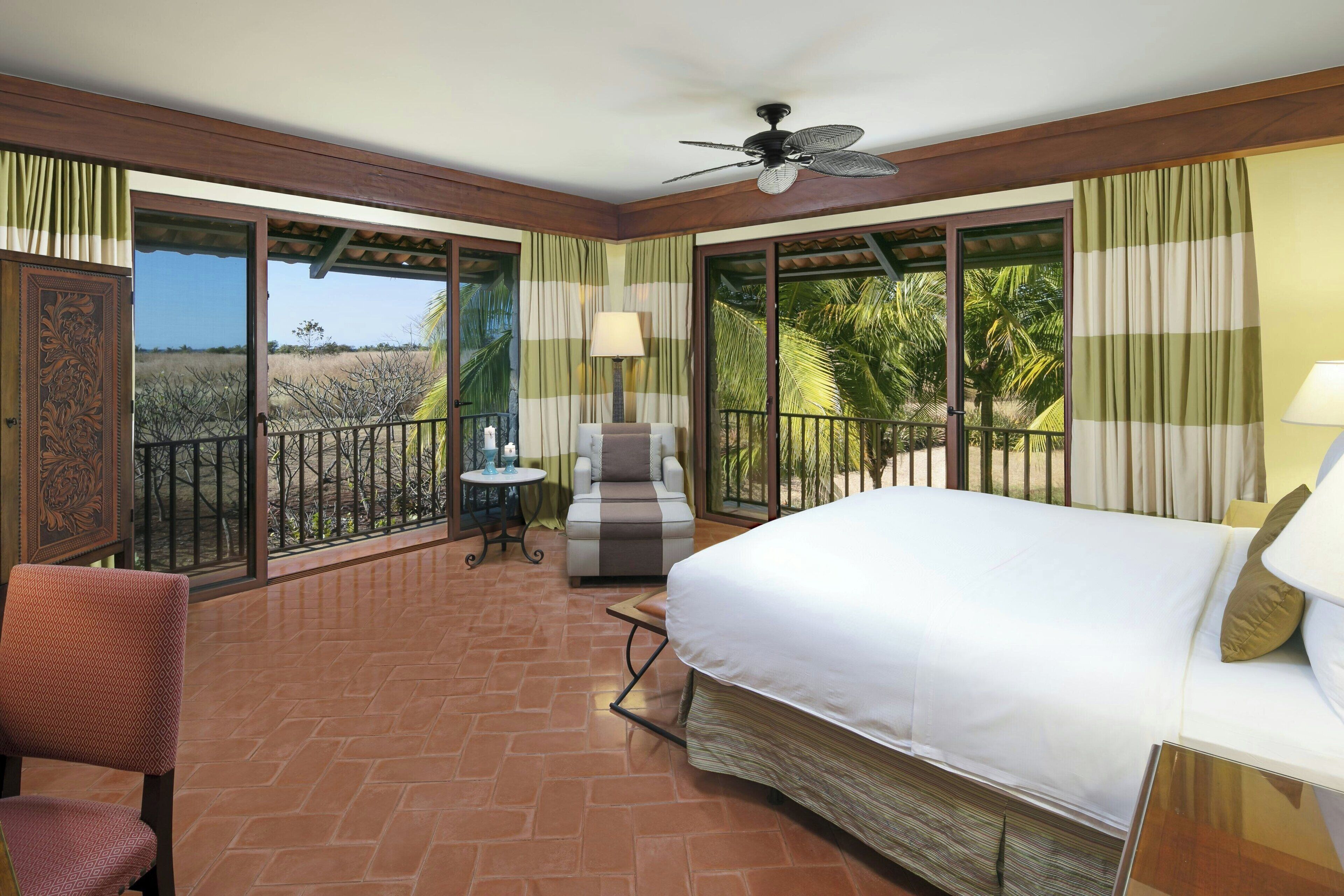 A hotel room with a large bed, a chair, a small table, and a balcony with a view of palm trees.