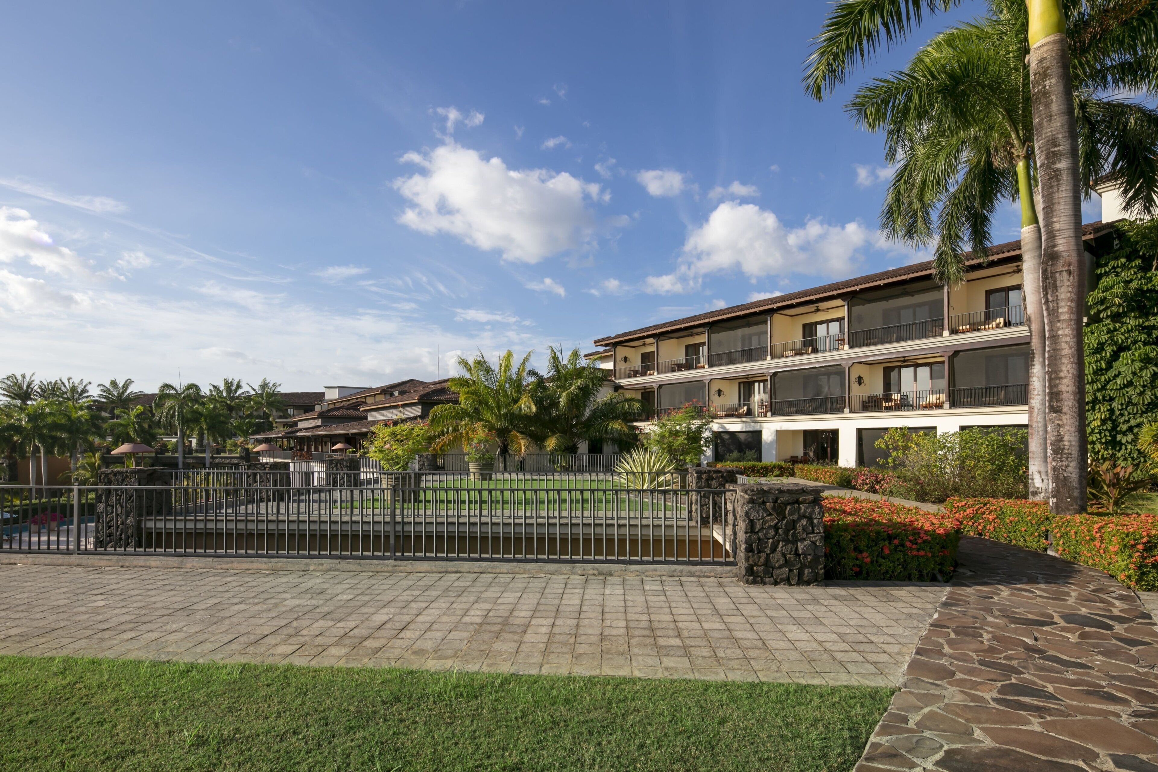 Un hotel de varios pisos con un patio empedrado de piedra, palmeras y un área cercada.