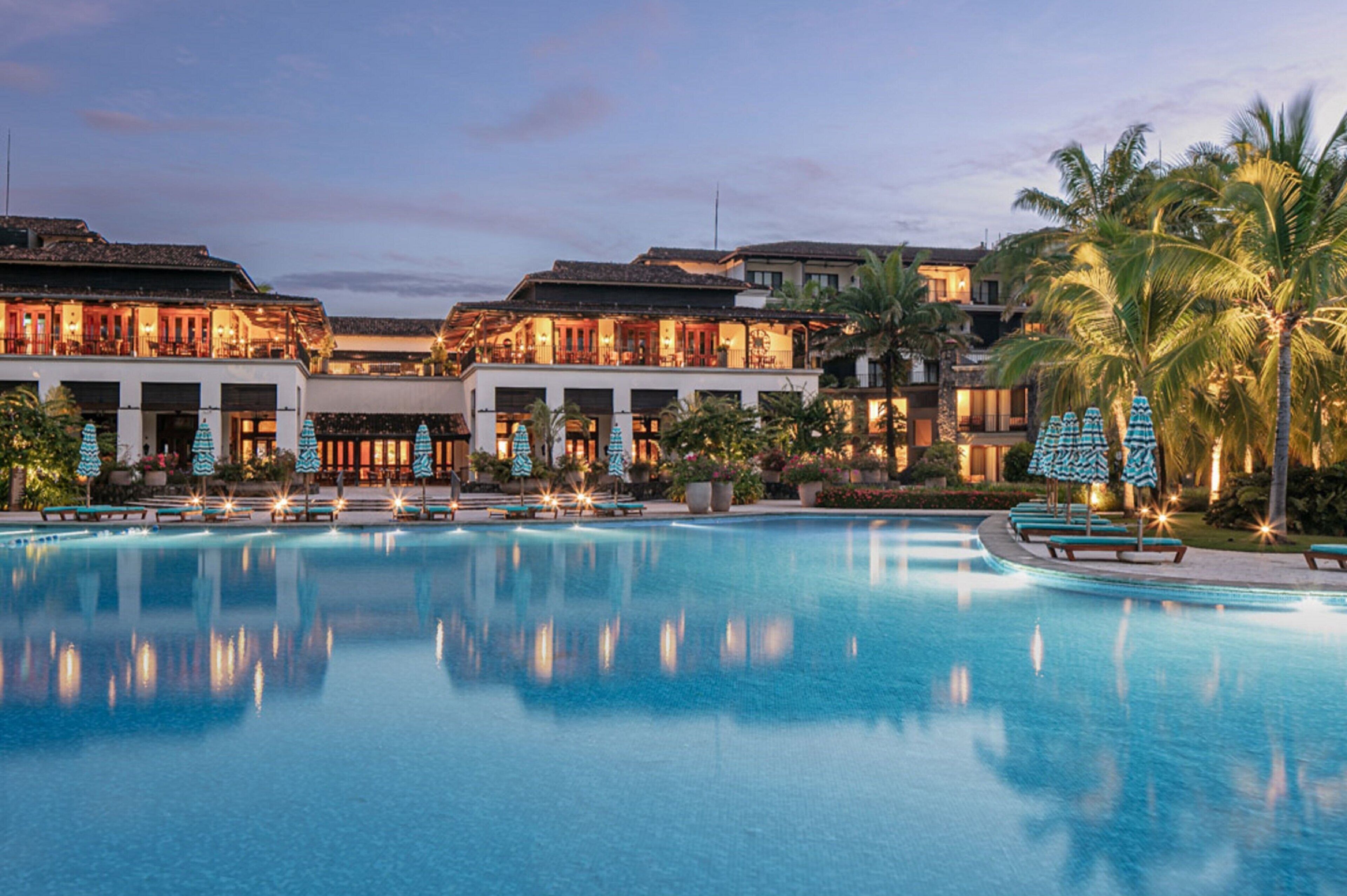 A resort with a large swimming pool, multiple buildings with balconies, and palm trees.