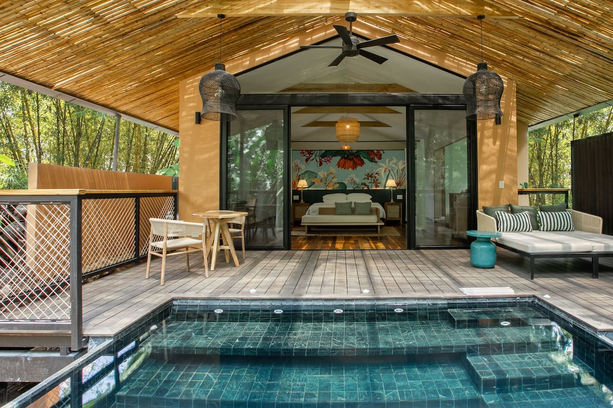 A modern outdoor pool area with a wooden deck, a glass-enclosed bedroom with a bed, and a seating area with a sofa and chairs.