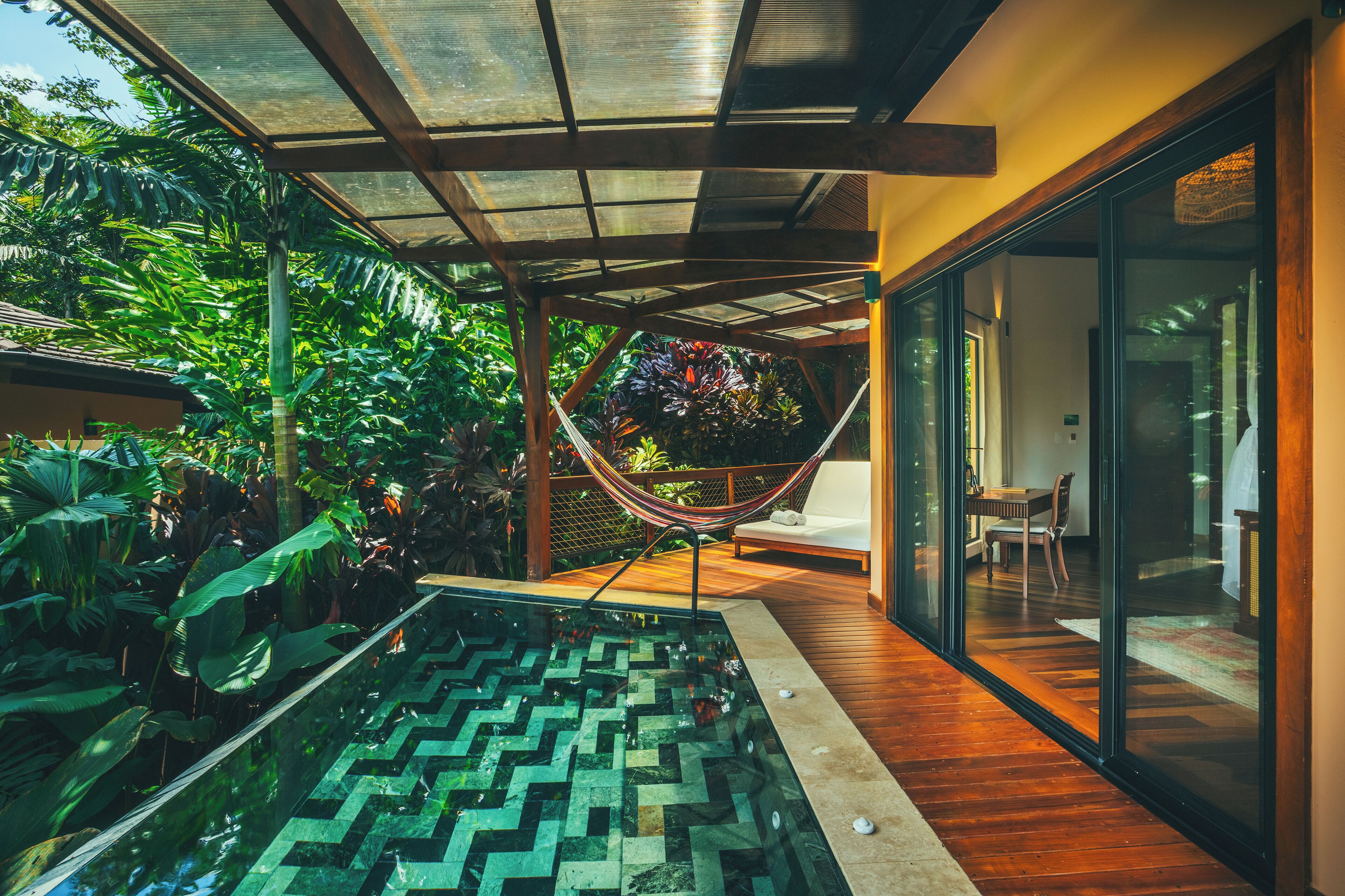 Uma piscina interior moderna com padrão geométrico de azulejos, rodeada por plantas tropicais exuberantes e uma rede.
