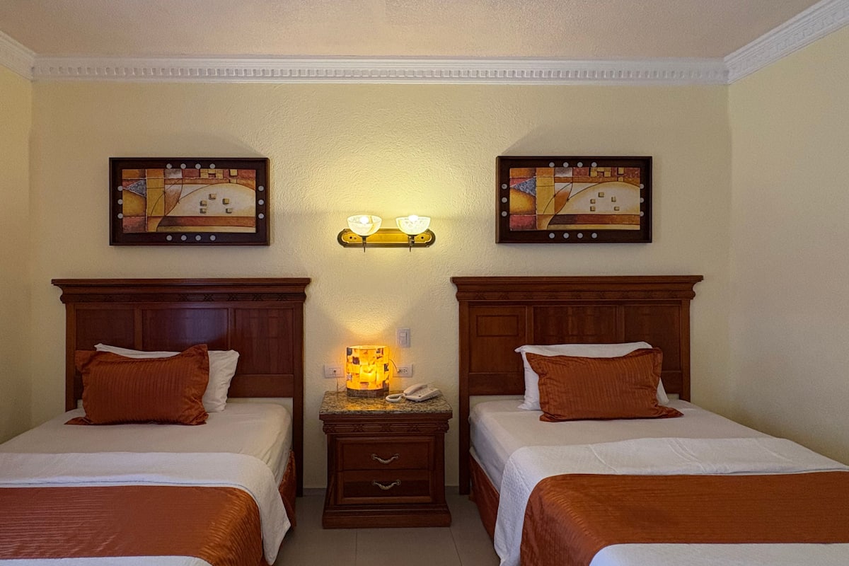 Une chambre d’hôtel avec deux lits, une table de chevet, une lampe et deux tableaux encadrés au mur.