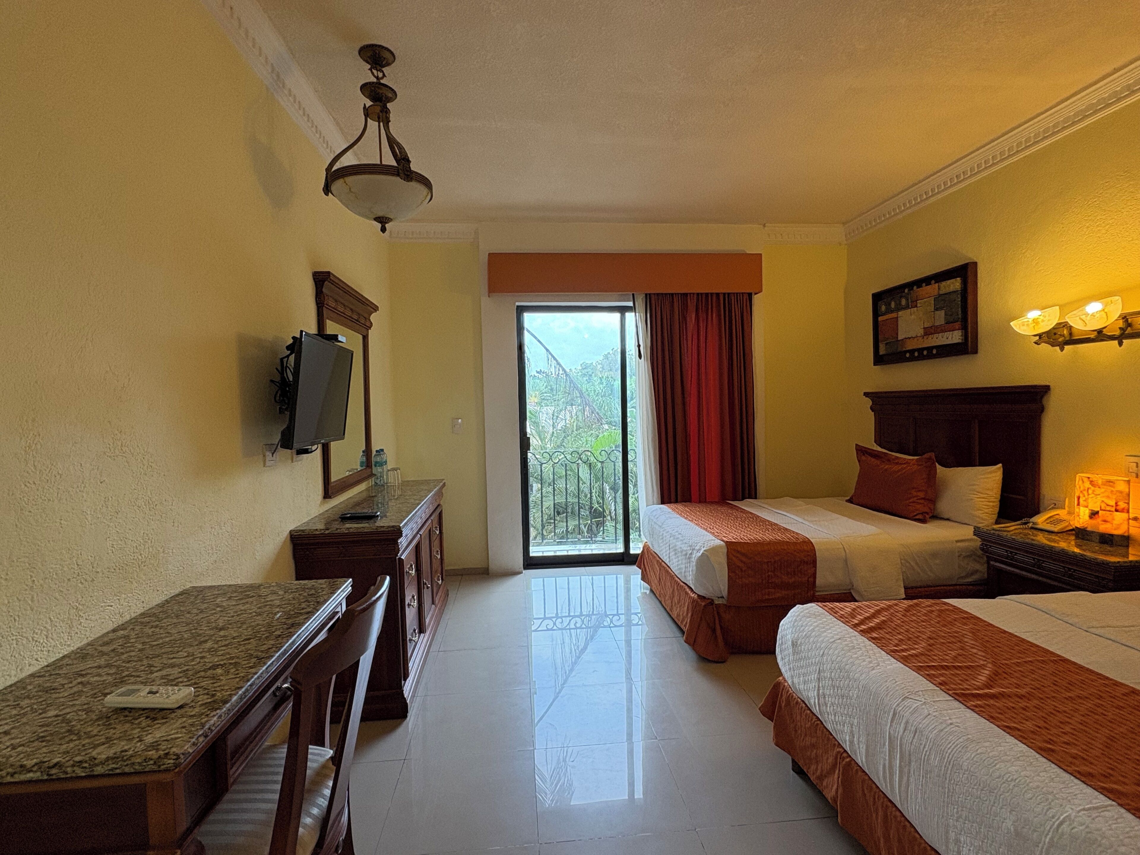 A hotel room with two beds, a desk, a TV, and a balcony with a view.