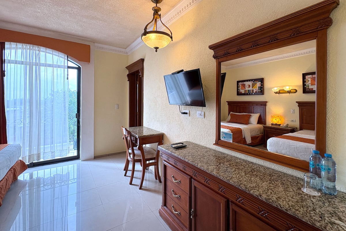 Une chambre d’hôtel avec un grand lit, un téléviseur fixé au mur, un coin repas avec une table et des chaises, et une grande fenêtre avec des rideaux.