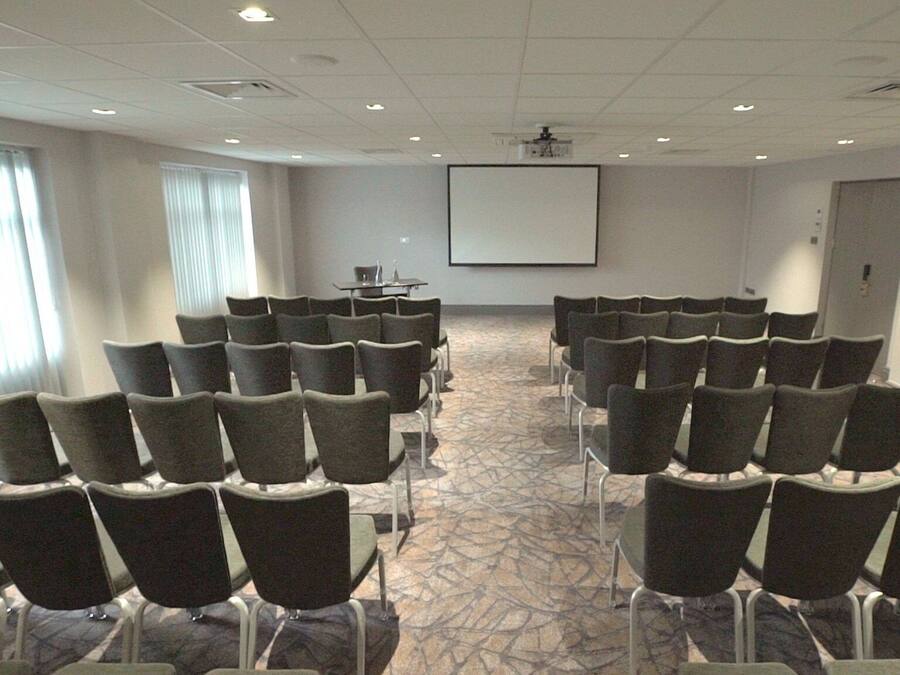 Un salón de conferencias con filas de sillas orientadas hacia una pantalla de proyección, ventanas amplias con persianas y suelo alfombrado.
