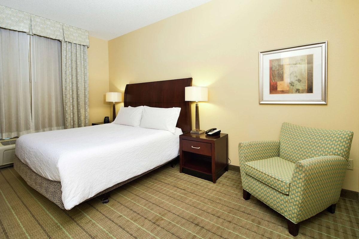 A hotel room with a bed, a nightstand with a lamp, a chair, and a framed picture on the wall.
