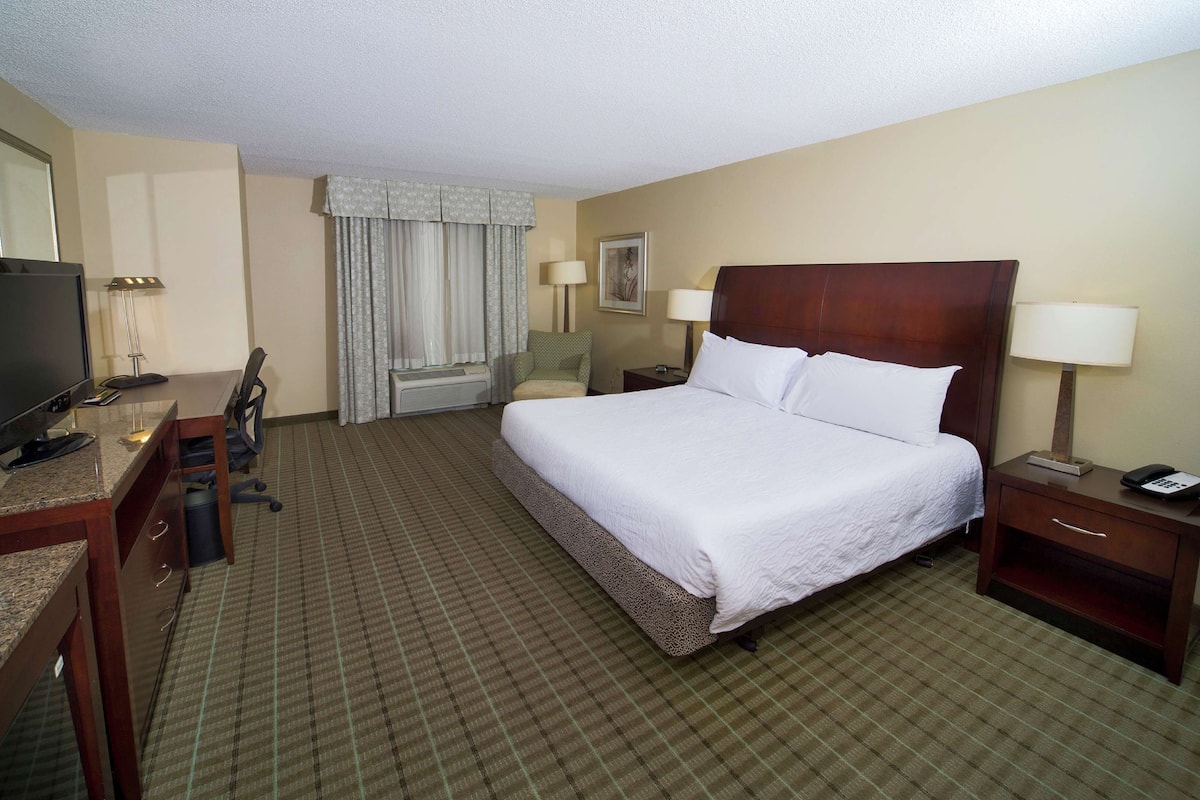 A hotel room with a bed, a desk with a TV, a chair, a nightstand with a phone, and a window with curtains.