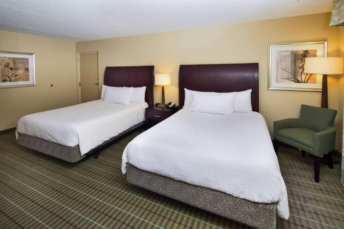 A hotel room with two beds, a green chair, and a lamp.
