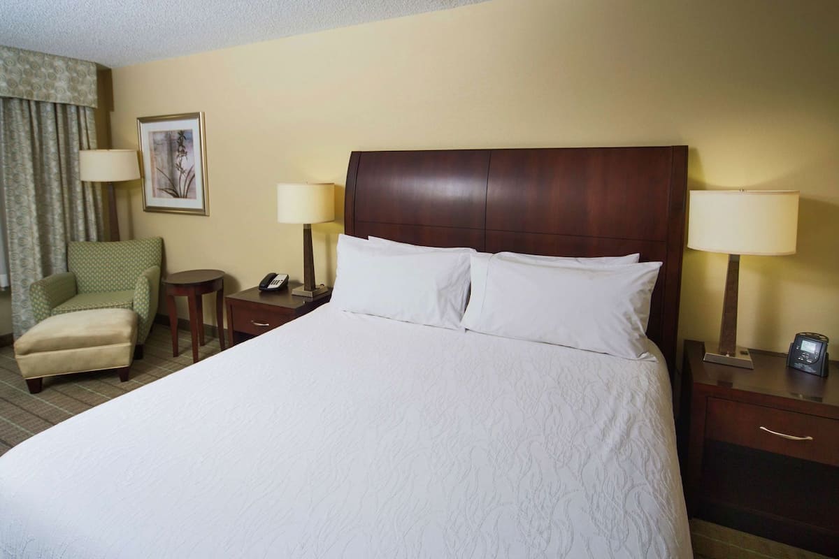A hotel room with a large bed, two bedside tables with lamps, a chair, a desk, a telephone, and a framed picture on the wall.
