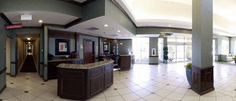 A hotel lobby with a reception desk, a hallway leading to rooms, and a large open space with a view of the outside.