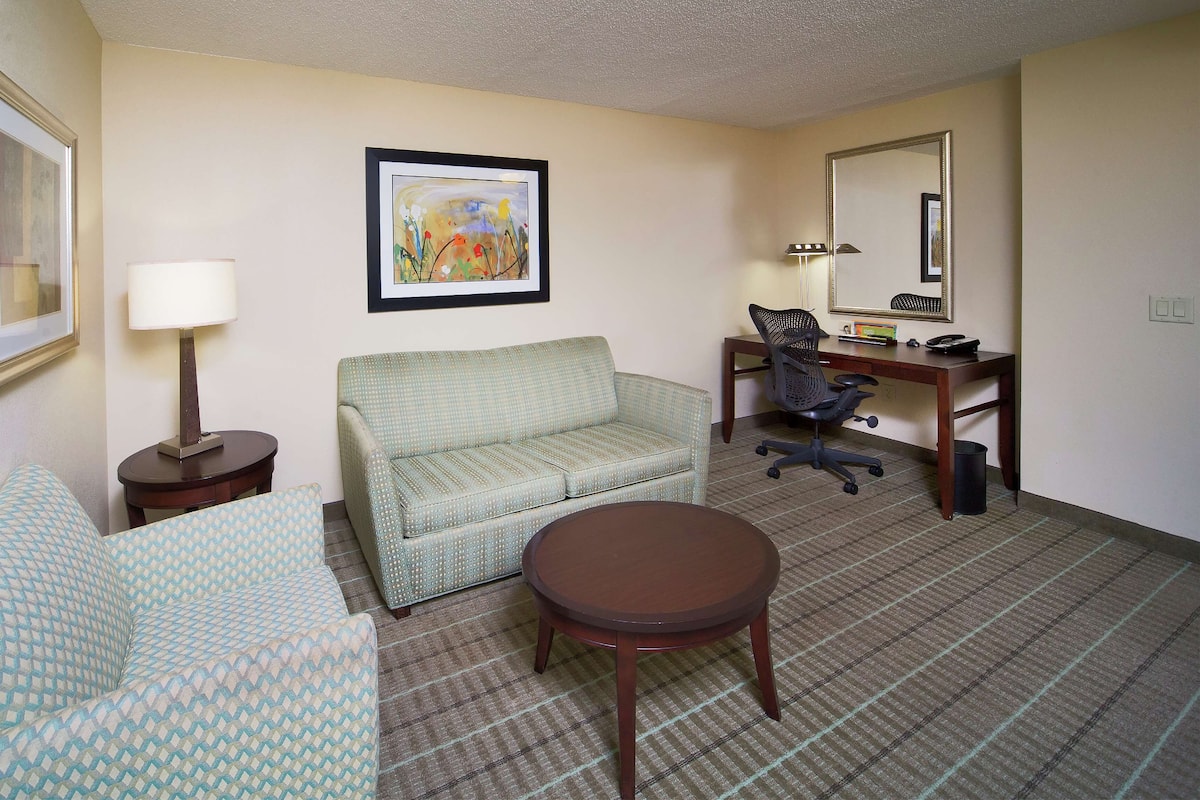 A hotel room with a sofa, a small round table, a desk with a chair, a lamp, and a framed picture on the wall.