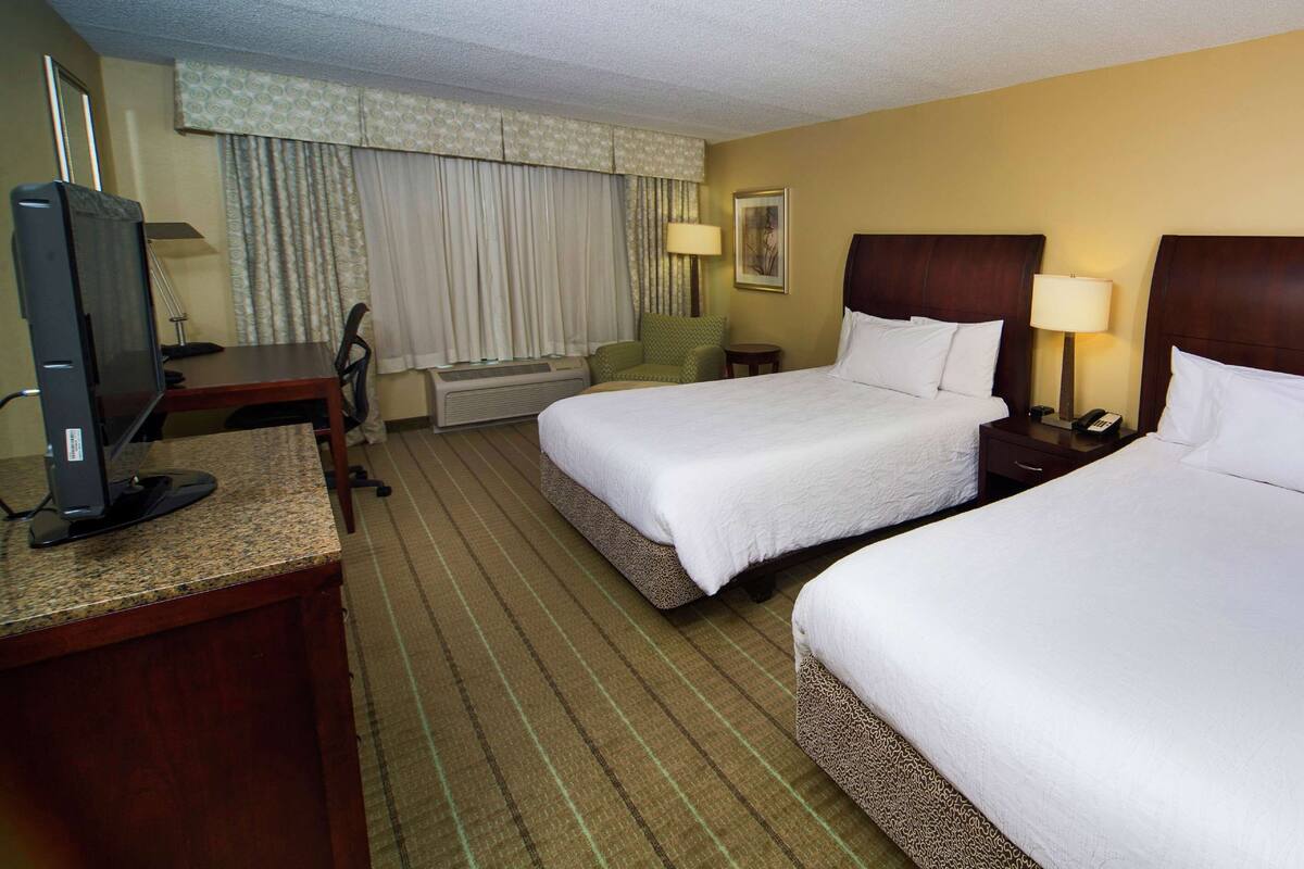 A hotel room with two beds, a desk with a computer, a chair, a lamp, and a window with curtains.
