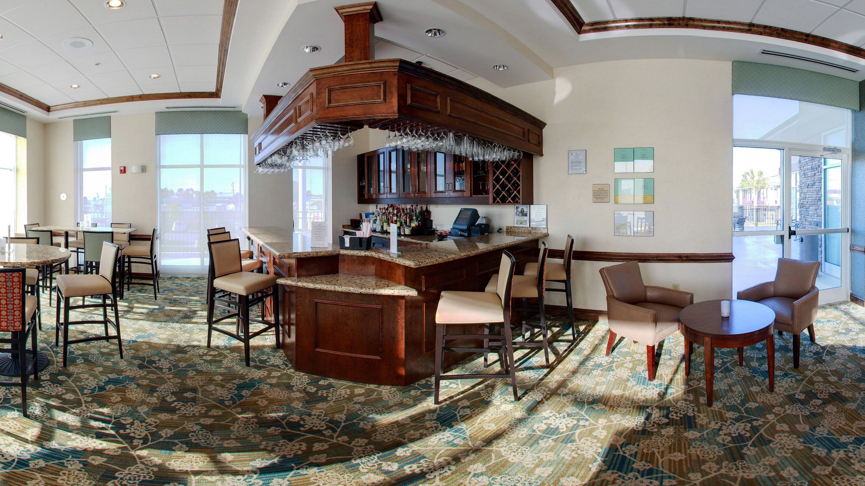 A spacious bar area with a wooden bar counter, multiple seating options including bar stools and chairs, and a patterned carpet.