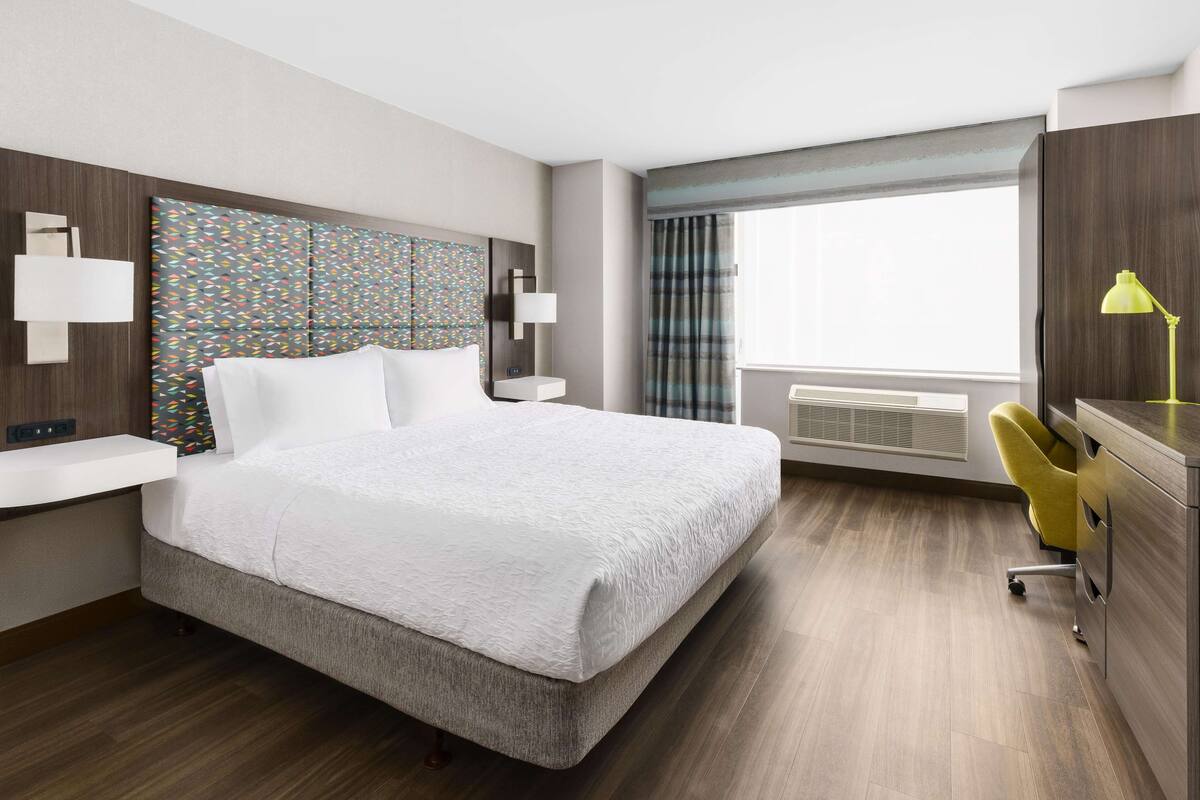 A modern hotel room with a large bed, a desk with a chair, a window with curtains, and a wall-mounted lamp.