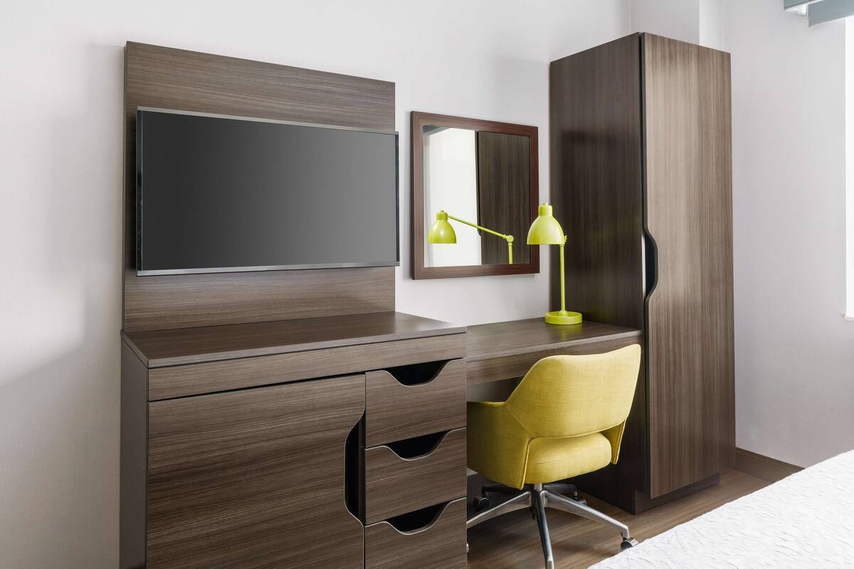 A modern bedroom with a wooden desk, a yellow chair, a flat-screen TV, a mirror, and a wardrobe.