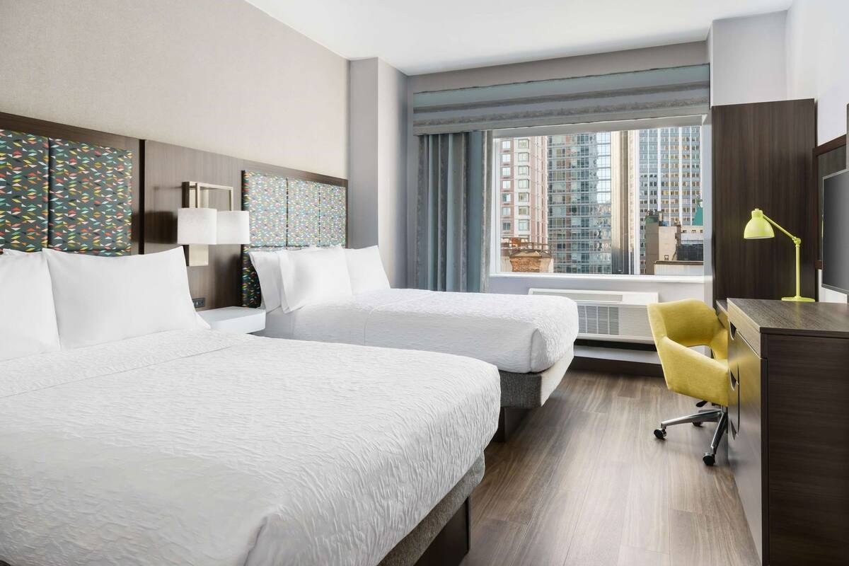A hotel room with two beds, a desk, a chair, a TV, and a window with a city view.
